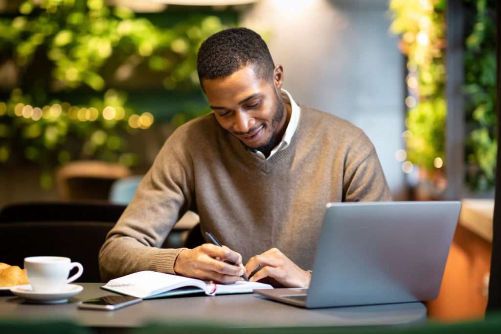 What is a Brain Dump & How Does it Help Beat Writer's Block. Man writes in journal in front of his computer.